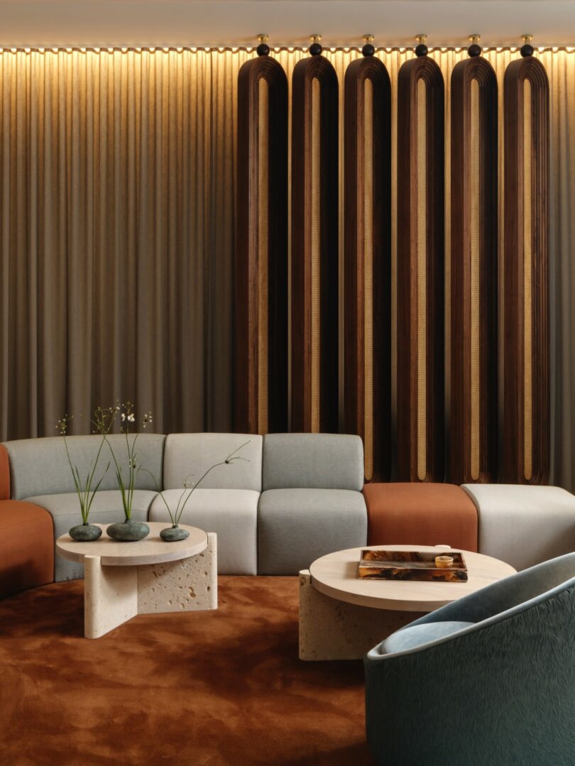 Modern living room with a curved sectional sofa in shades of brown and gray, stone coffee tables, and vertical wooden wall accents under soft ambient lighting. Elegant and minimalistic decor.