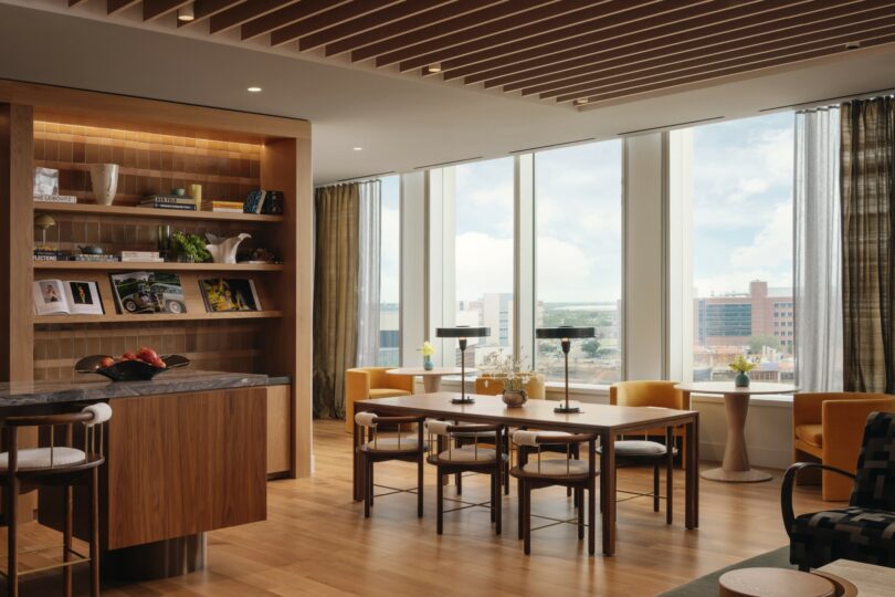 A modern room with floor-to-ceiling windows, wooden furniture, and shelves with books and decor. Light streams in, illuminating a long dining table with chairs and a countertop with stools.