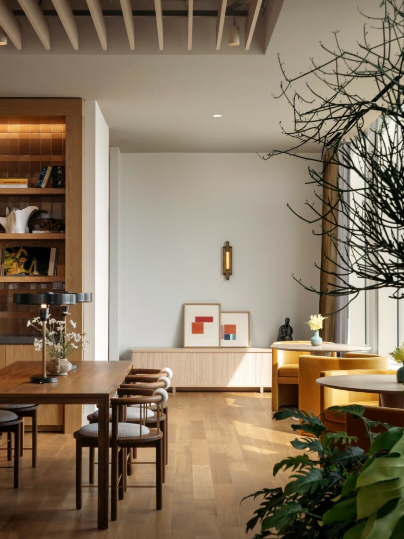 A modern living and dining area with wooden flooring, contemporary furniture, minimalistic decor, and a large window. Indoor plants add greenery to the space. Two framed artworks are displayed on a sideboard.