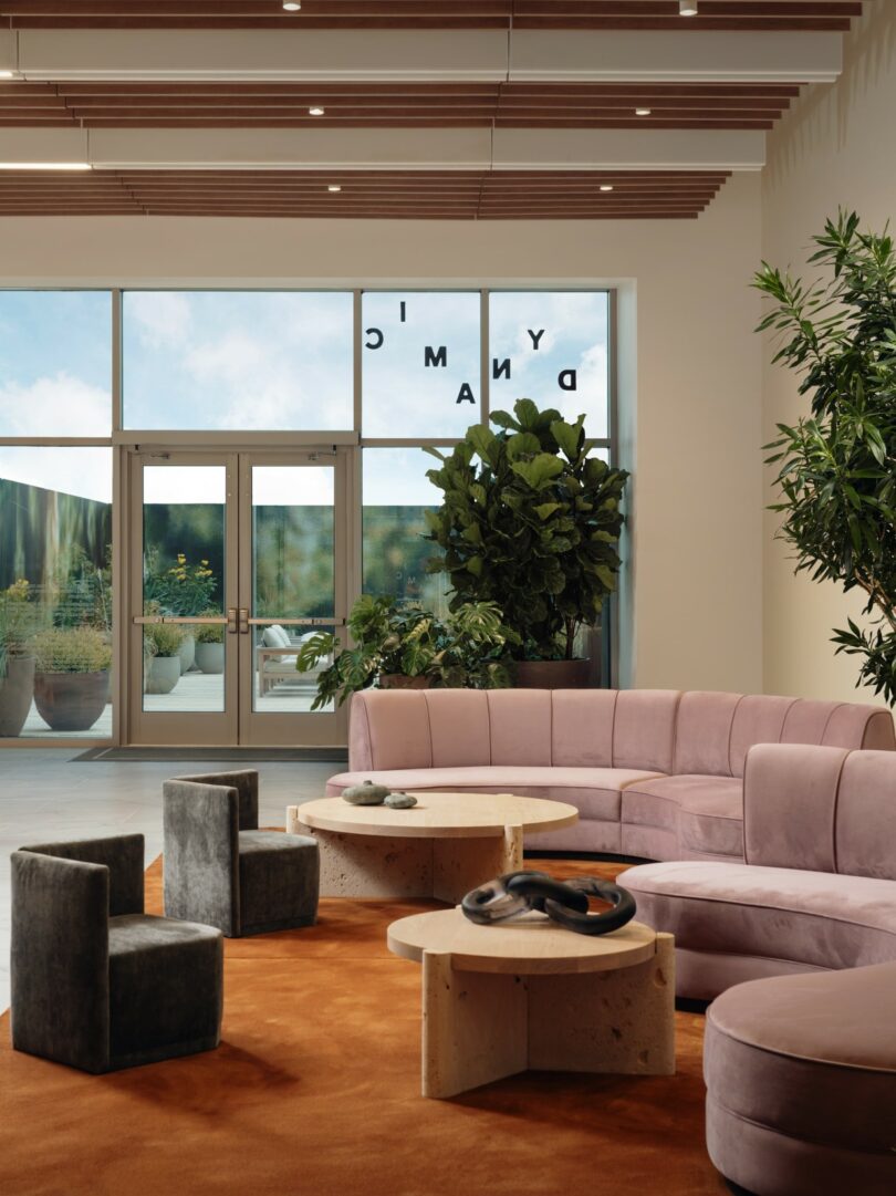 A modern lobby with a curved pink sofa, round stone tables, dark upholstered chairs, large indoor plants, and glass doors with partial text visible, leading to an outdoor area.