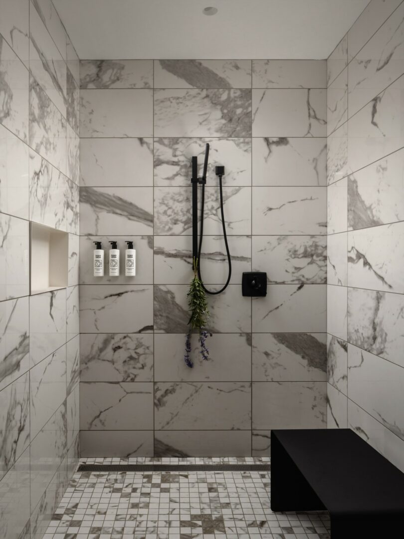 A modern shower with white and grey marble tile, a black hand-held showerhead, three bottles on a built-in shelf, and a black bench. A bundle of fragrant herbs hangs from the showerhead.