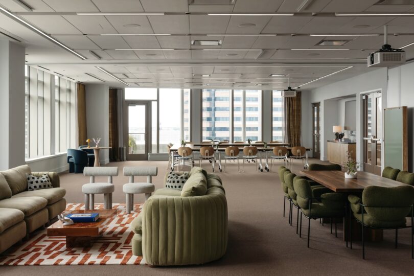 A modern office lounge with green sofas, a wooden table with chairs, and a conference area in the background, featuring large windows and neutral tones.