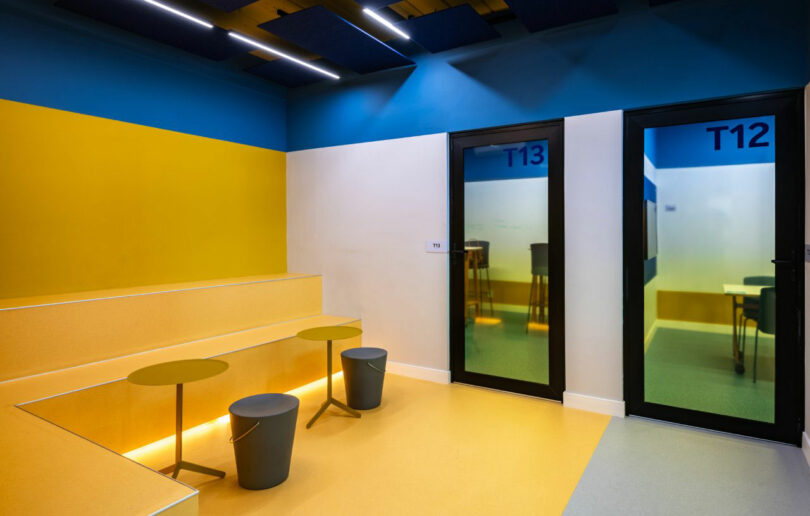 A modern interior by Estúdio Protobox features tiered yellow seating, small circular tables, and two glass doors labeled T12 and T13, with rooms containing tables and chairs visible through the doors.