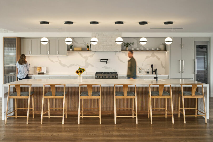 Modern kitchen designed by IA Interior Architects, featuring a long island with wooden stools and hanging pendant lights. Two people interact with appliances on opposite sides of the room, amidst white cabinetry and a subway tile backsplash.