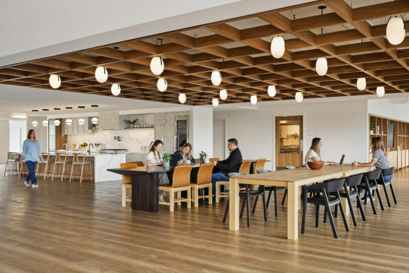 Open office space designed by IA Interior Architects, featuring wooden ceiling and floors, modern lighting, and a shared kitchen. Several people are sitting at long tables, working on laptops and conversing. One person stands near the kitchen.