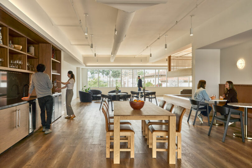 A communal space by IA Interior Architects, where people engage in various activities, including using the kitchen, sitting at a large wooden table, and conversing at smaller tables. The area features modern furnishings and ample natural light.