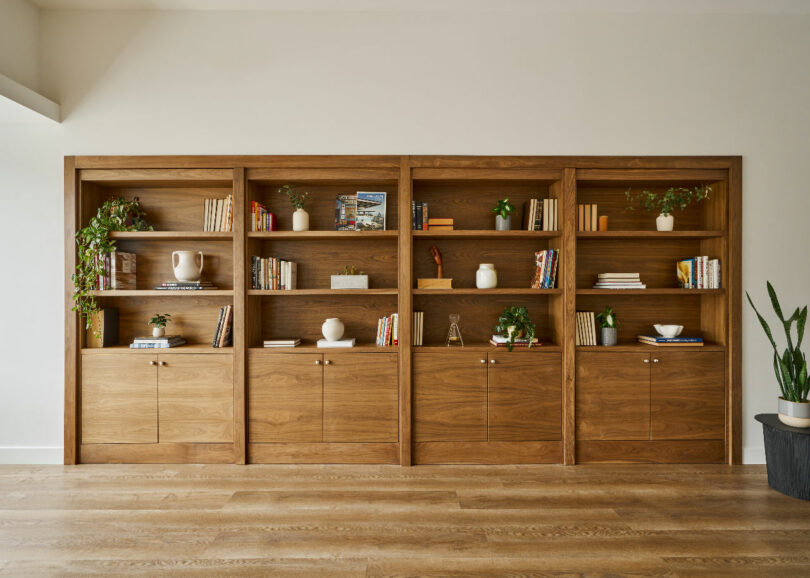 A wall with four wooden bookcases, each filled with books, decorative items, and potted plants, stands on a wooden floor. Designed by IA Interior Architects, this setup marries functionality and aesthetic appeal harmoniously.