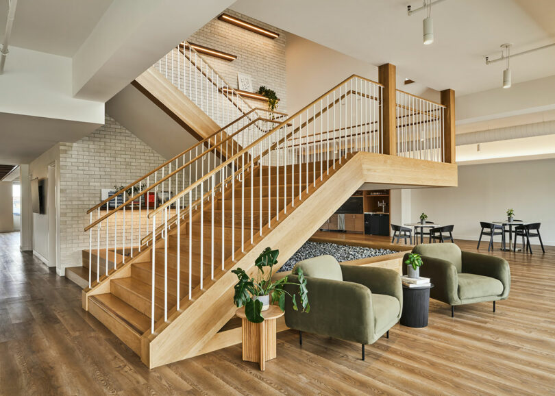 A modern interior by IA Interior Architects features a wooden staircase, green chairs, small plants, and tables with chairs. The space embraces both natural light and artificial lighting, showcasing a clean and minimalistic design.