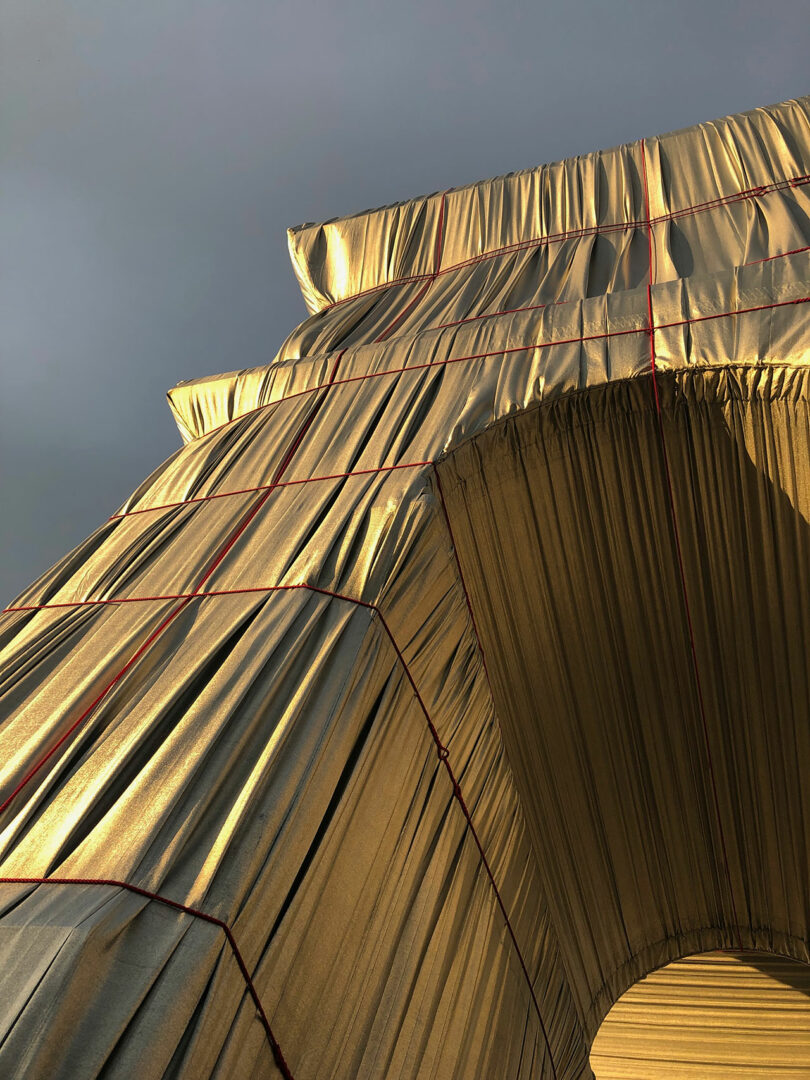 Close-up of a large structure draped in silver fabric with red ropes, set against a gray sky. The fabric appears to be tightly wrapped, creating pleats and folds.