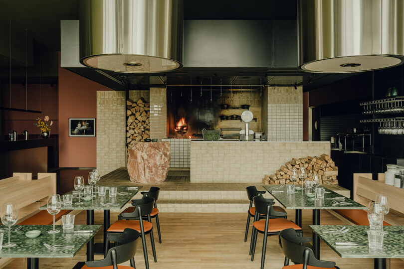 A modern restaurant interior featuring green marble tables with wooden chairs, an open kitchen with a wood-fired oven, stacked firewood, and two large cylindrical exhaust hoods above.