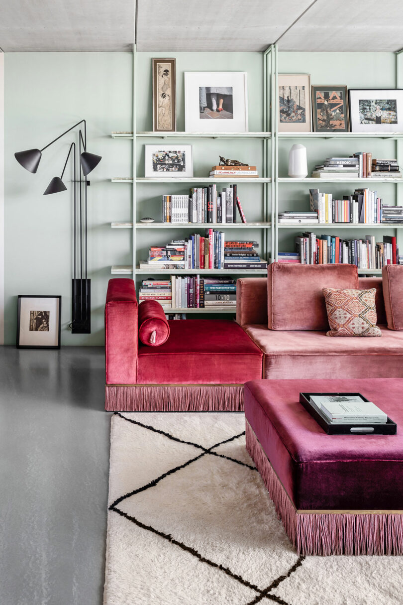 A modern living room with a red velvet sectional sofa, a matching ottoman, a white and black rug, a black floor lamp, and a bookshelf filled with books and framed art.