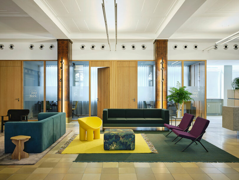 Modern office lounge area with colorful furniture, including green and yellow sofas, purple chairs, and a patterned ottoman on rugs. Glass-walled meeting rooms are visible in the background.