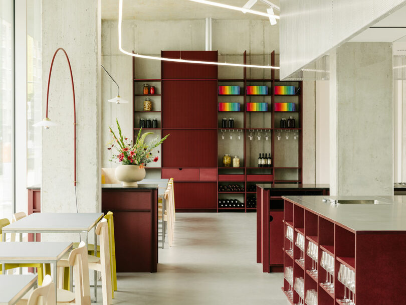 Modern kitchen with red cabinets, colorful books, wine bottles, and a vase of flowers on the counter. Tables and chairs are placed nearby, with a contemporary lighting fixture overhead.