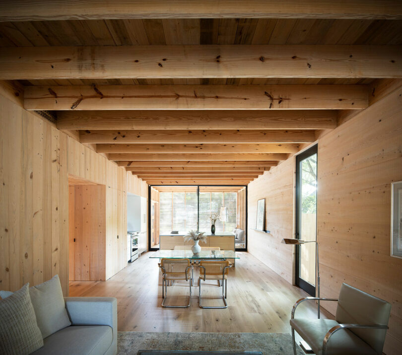 A living room with wooden walls and ceiling beams, featuring a sofa, a glass dining table with four chairs, and large windows letting in natural light.