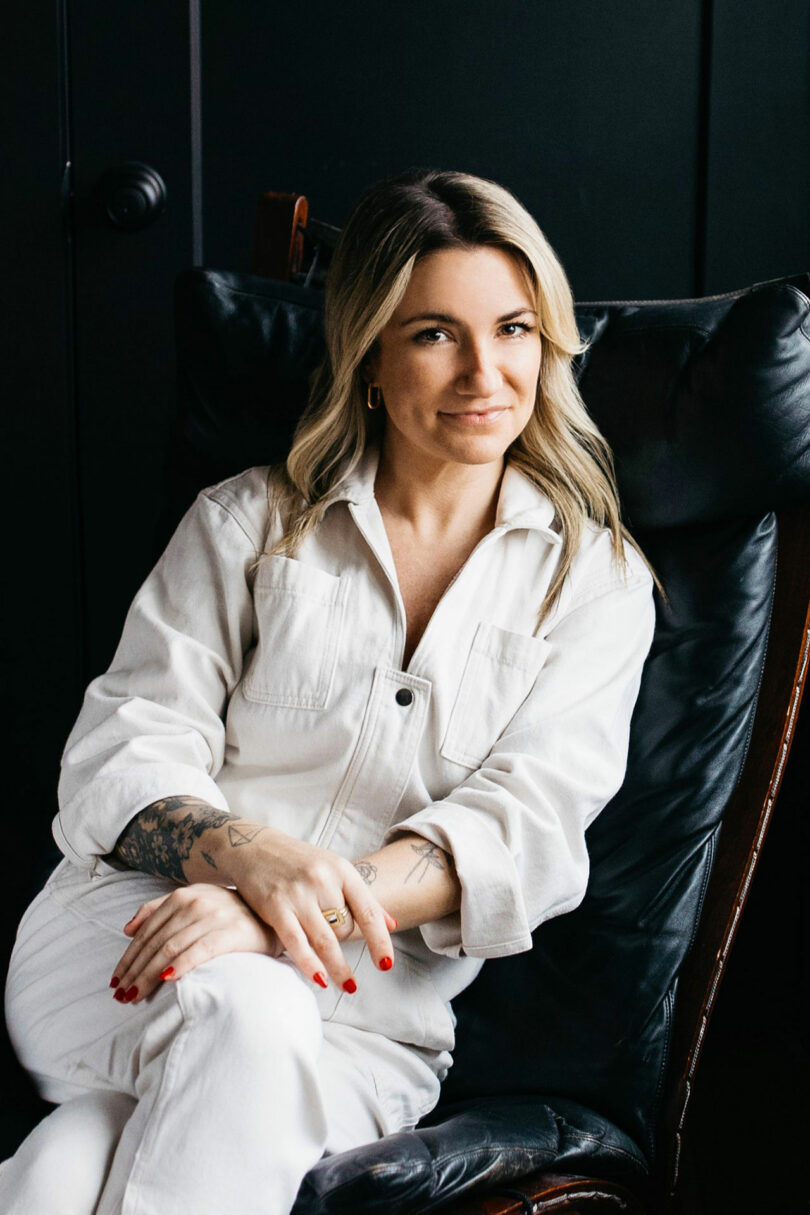 Kate Rohrer, a woman with blonde hair, sits in a black leather chair wearing a white button-up outfit. She has tattoos on her forearms and is smiling slightly.