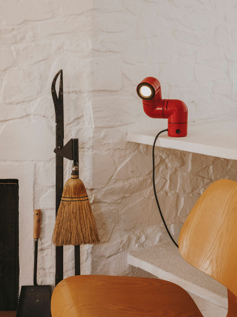 A red desk lamp sits on a white tabletop with wooden chairs nearby, a broom, and a fireplace tool set against a white brick wall in the background, evoking the clean and modern aesthetic.