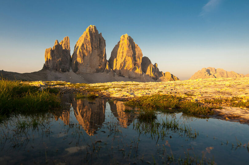 A scenic view of mountain peaks reflected in a small foreground puddle on a clear day. The landscape features rocky terrain and green grass under a blue sky.