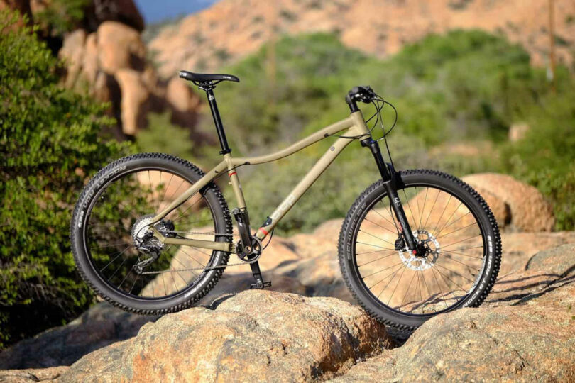 A mountain bike with a green frame is positioned on rocky terrain outdoors, surrounded by greenery and rocky formations in the background.