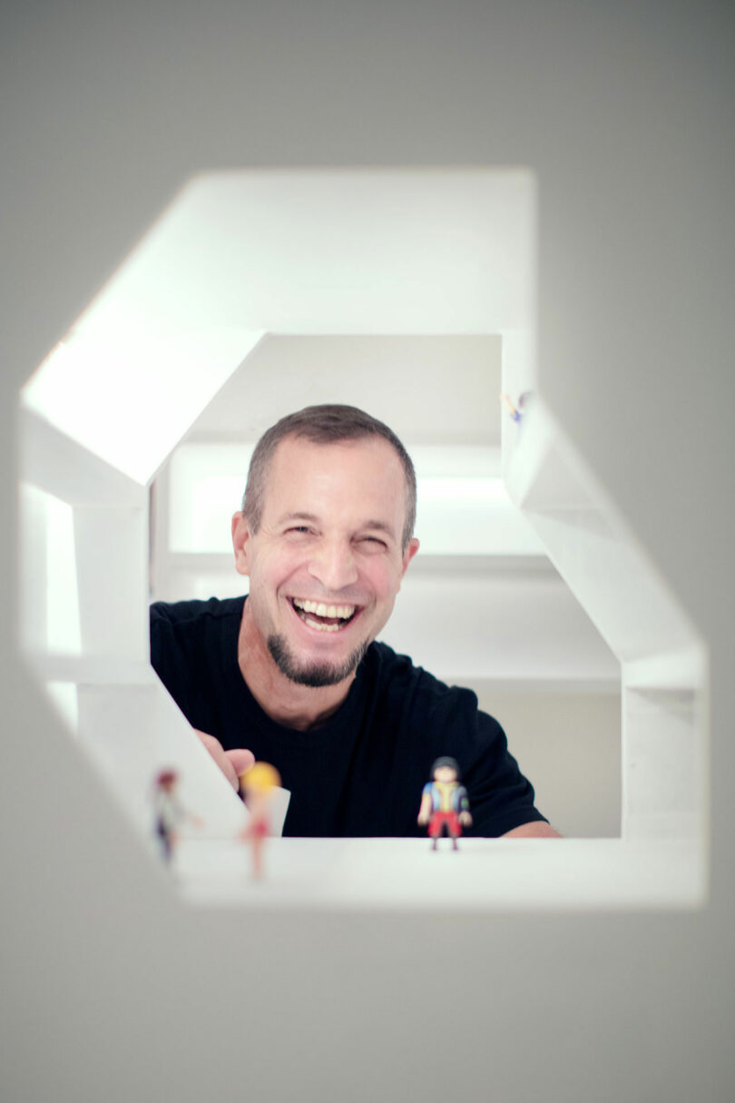 A person with a beard, wearing a black shirt, smiles while looking through a hexagonal white opening. Several small toy figures are visible in the foreground.