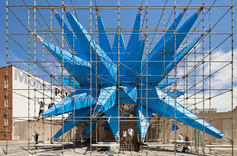 Workers assemble a large blue spiked structure within scaffolding against a clear sky.