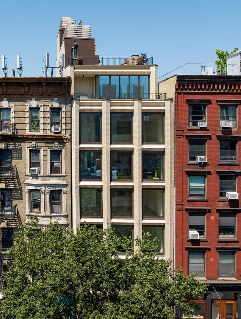 A modern, multi-story building with large windows is situated between two older, brick buildings in an urban setting. Trees partially obscure the lower part of the buildings.