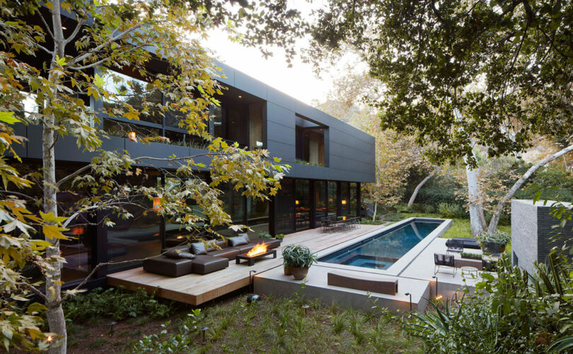 Modern two-story house with large windows, surrounded by trees, featuring an outdoor deck, seating area, fire pit, and a rectangular swimming pool. Designed with the signature touch of Ron Radziner.