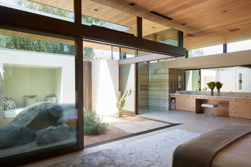 A modern, minimalist bedroom designed by Ron Radziner features large windows, a wooden ceiling, and an indoor garden with a cactus. Built-in cabinetry and a cozy seating nook enhance the space, while natural light beautifully illuminates every corner.