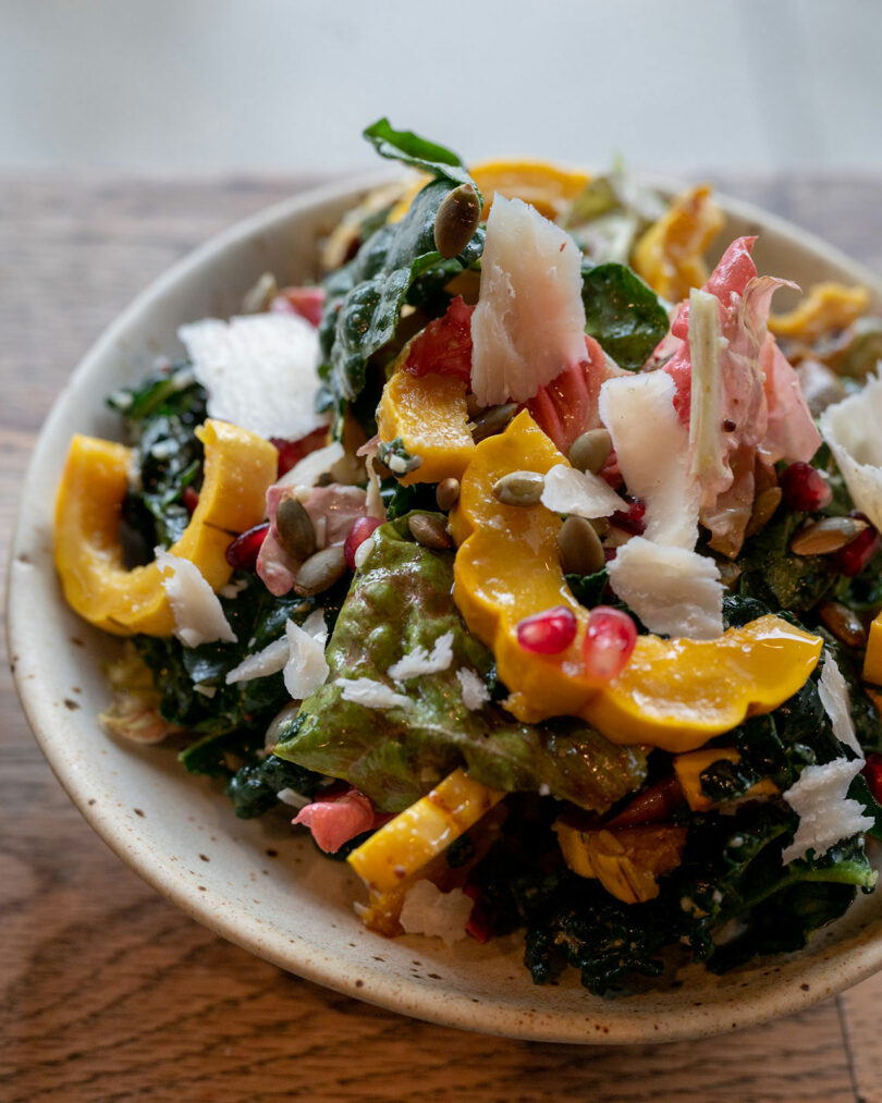 A bowl of salad with mixed greens, shaved cheese, pumpkin seeds, pomegranate seeds, and roasted yellow squash rests on a wooden surface.