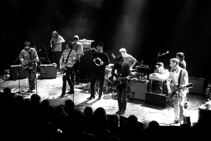 Black and white image of a band performing on stage with multiple musicians playing instruments and a crowd.