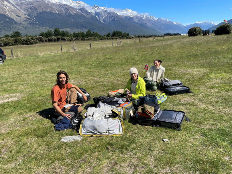 Three people are sitting on the grass in an open field with mountains in the background, organizing items from open suitcases around them.