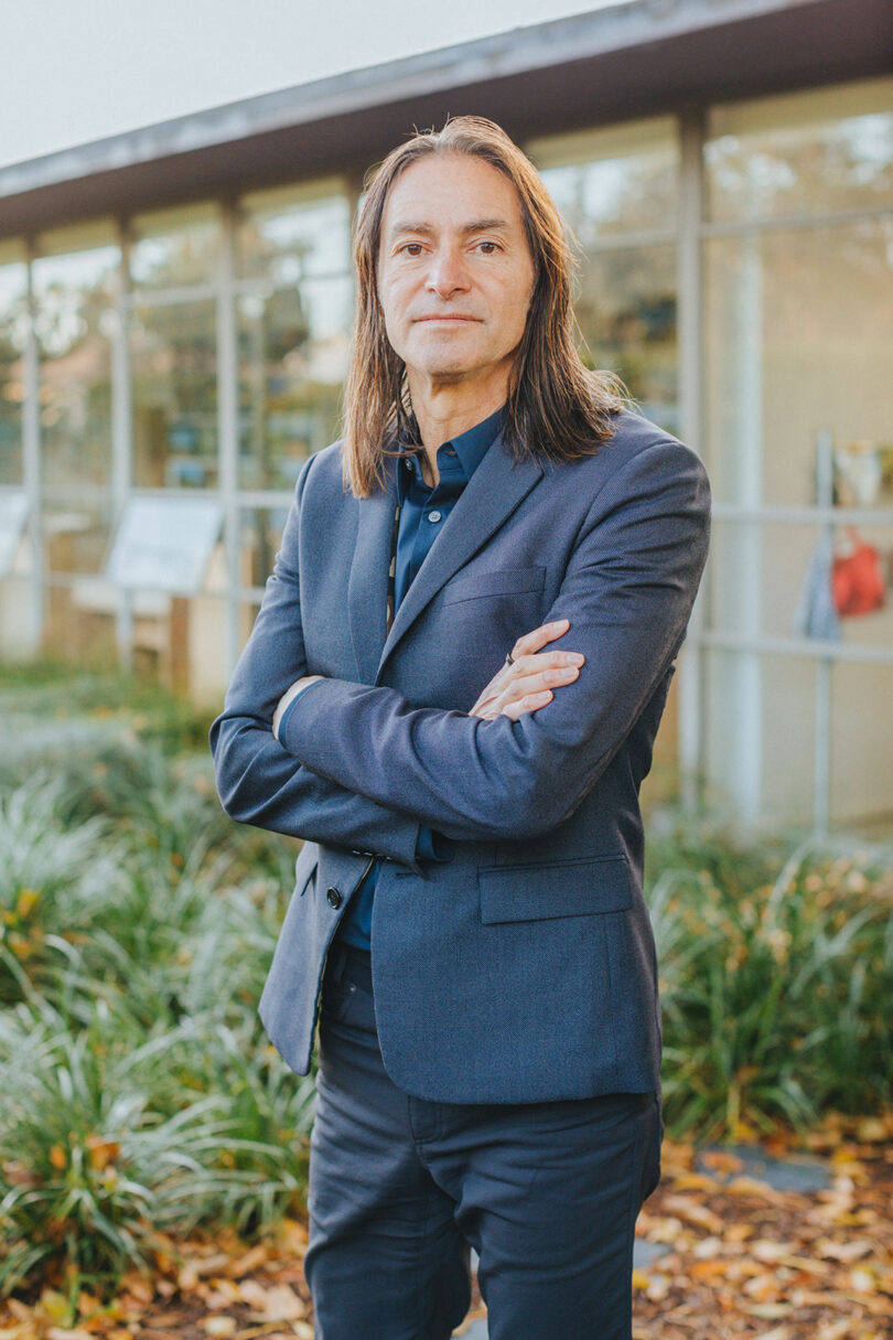 A person with long hair stands outdoors with arms crossed, wearing a dark blazer and blue shirt. A building with large glass windows and greenery.