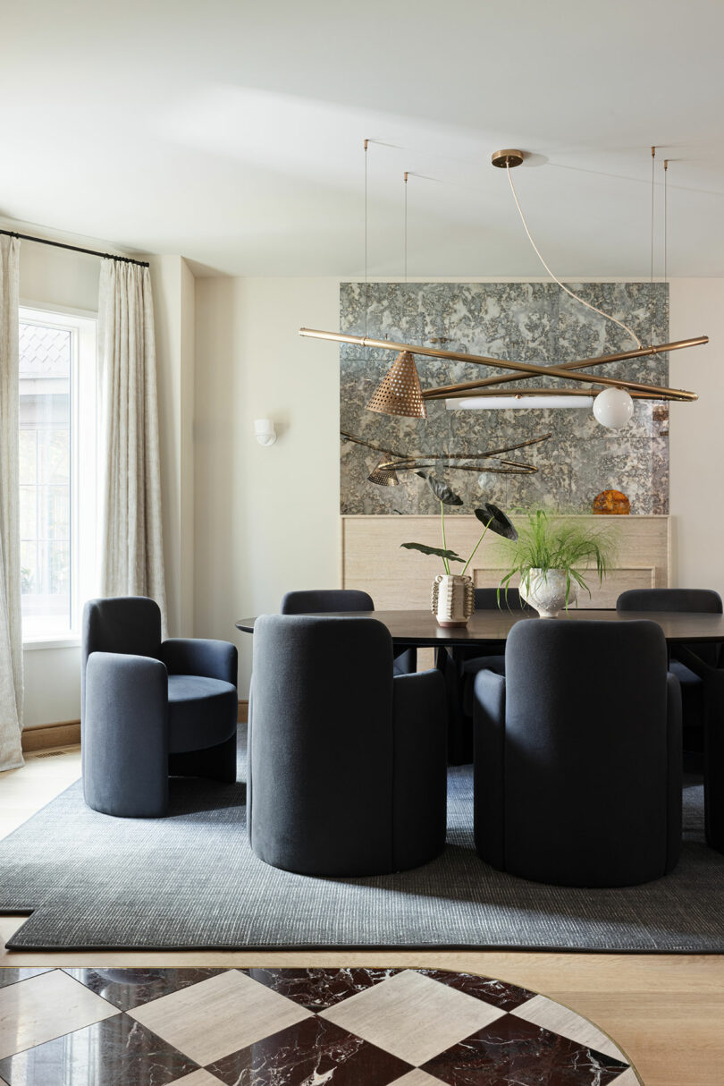 A modern dining room with round black chairs, a rectangular table, contemporary light fixtures, a textured wall, and large windows with curtains, all set on a geometric patterned rug.