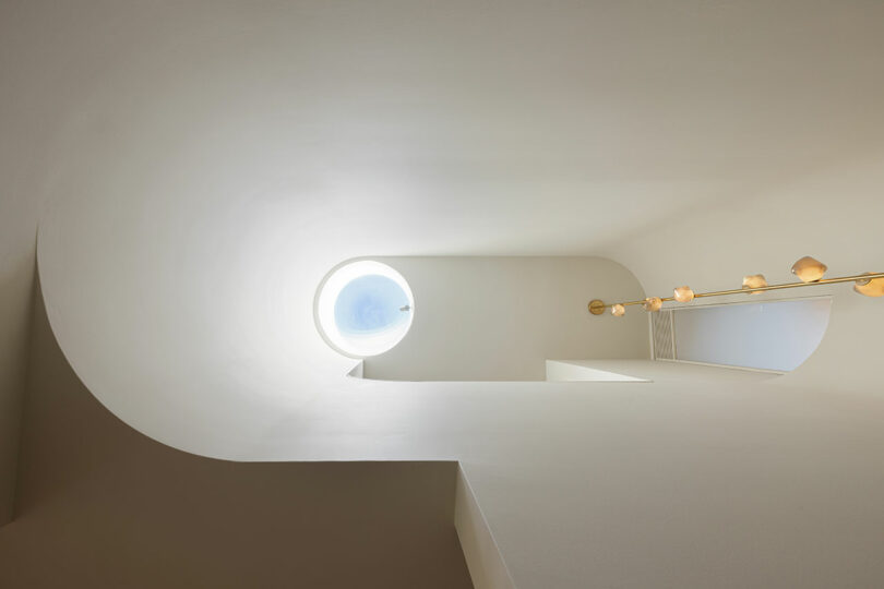 A staircase is shown from below, with curving white walls and a round window at the top letting in light. A line of spherical lights is mounted on the right wall.