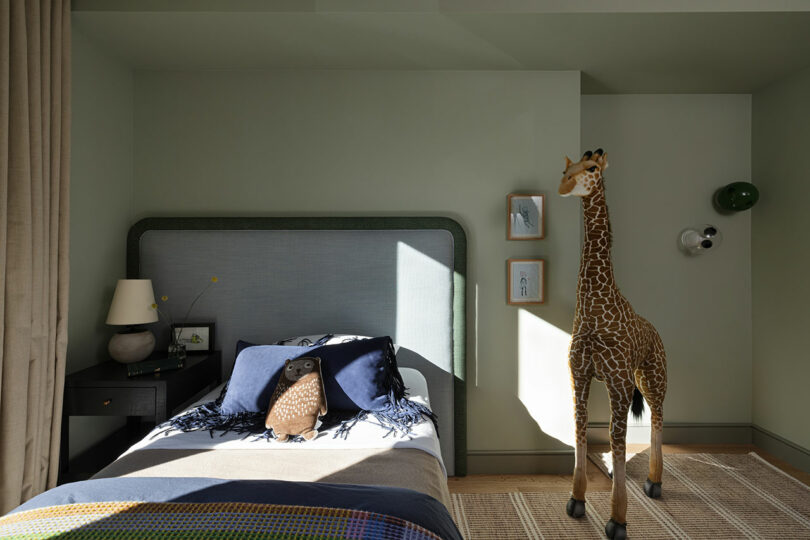 A bedroom with a neatly made bed featuring blue and gray bedding, a black nightstand with a lamp, framed artwork on the wall, and a large toy giraffe standing on the floor beside the bed.