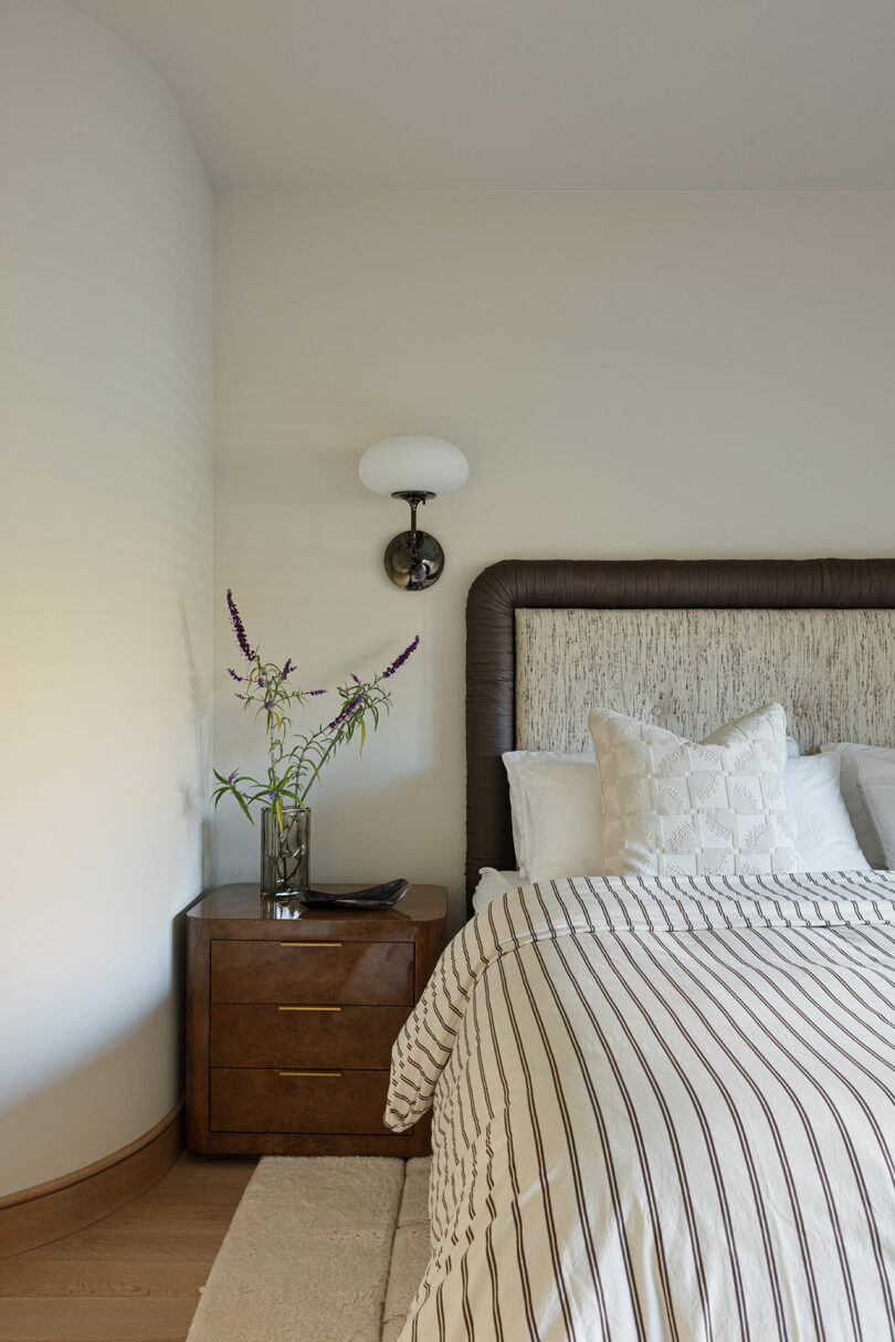 A neatly made bed with striped bedding is next to a wooden nightstand with two drawers. The nightstand has a vase of flowers, a book, and is lit by a wall-mounted light fixture above it.