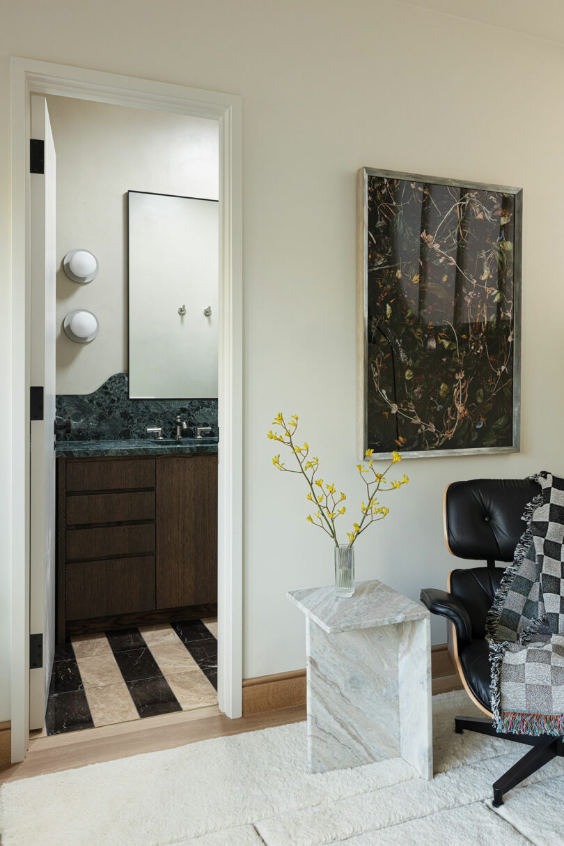 A small bathroom with dark wooden cabinets and green marble countertop is visible through an open door. The room in the foreground has a black modern chair, a white stone side table, and wall art.