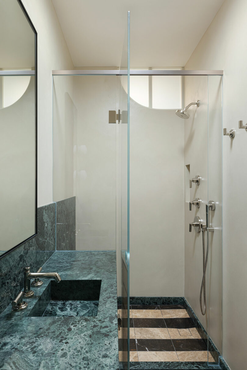 A modern bathroom features a green marble countertop with a sink, adjacent to a walk-in glass shower with a fixed showerhead and handheld sprayer, and a tiled floor.