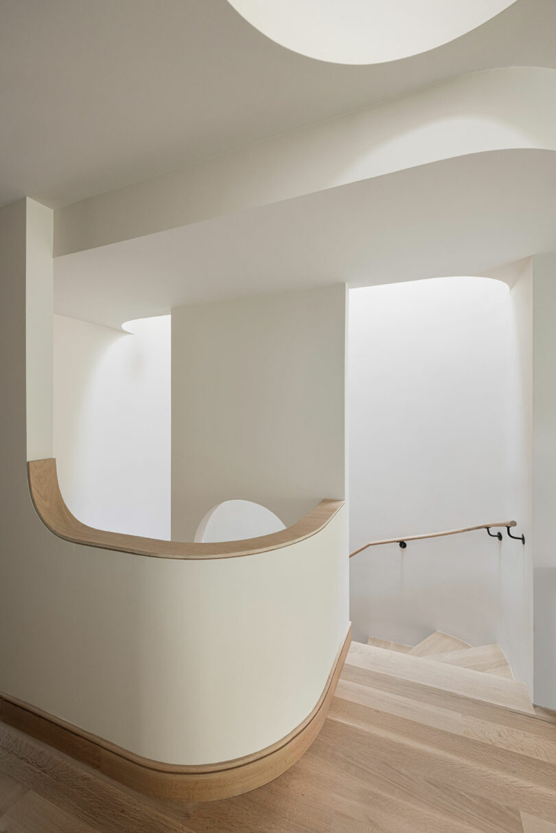 A minimalist, light-filled staircase with white walls, wooden steps, and a curved wooden handrail. The design features clean lines and a soft color palette.