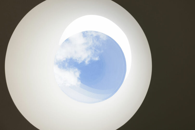 A circular skylight in a ceiling reveals a view of a blue sky with some clouds.