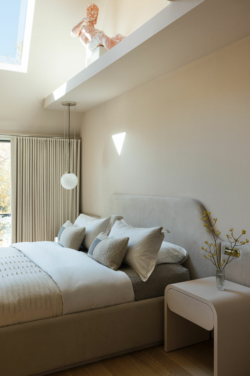 A modern bedroom with a neatly made bed, grey pillows, beige bedding, a small bedside table with a vase of flowers, and a glass pendant light. A skylight above displays a sculptural decoration.