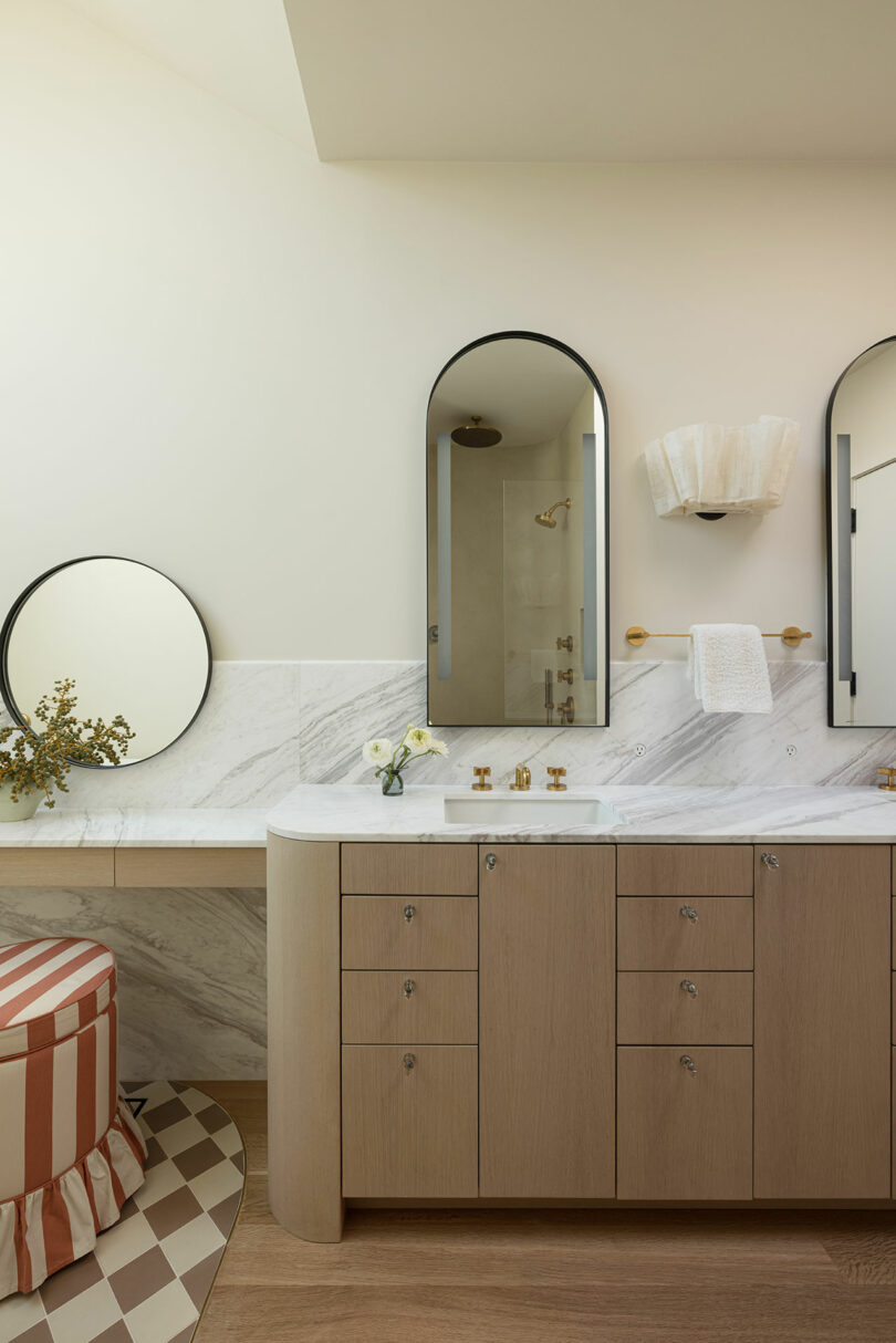 A modern bathroom features a dual-sink vanity with marble countertops, two oval mirrors, a wall-mounted light fixture, and a striped stool. A small round mirror and decorative greenery are on the side counter.