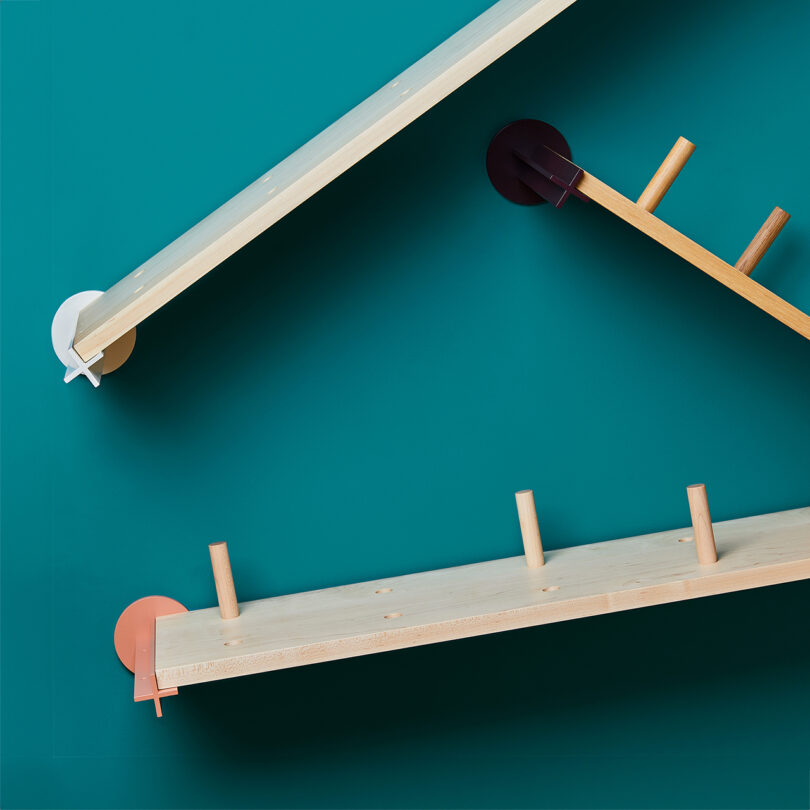 Three shelves with differently colored mounts are suspended diagonally against a teal background. Each ramp has wooden pegs inserted at intervals.