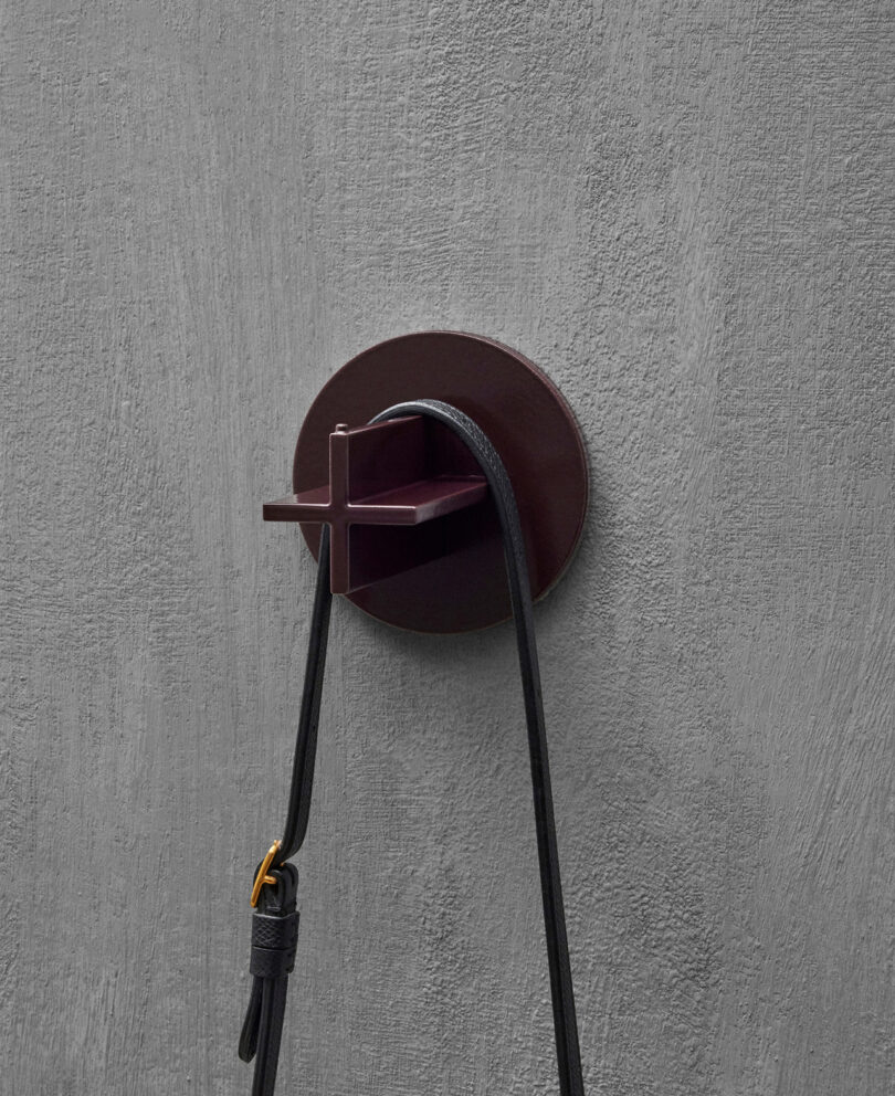 A burgundy wall hook shaped like a cross is mounted on a gray textured wall, holding a black strap.