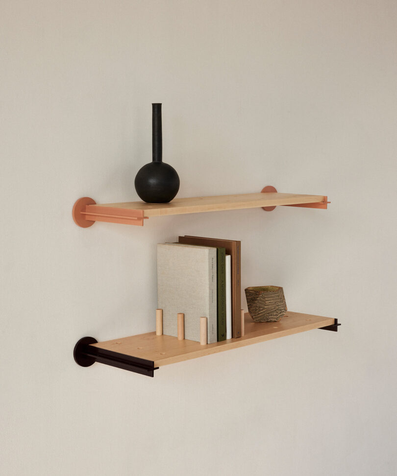 Two minimalist wall-mounted shelves: the top shelf holds a black vase, and the bottom shelf holds three books, a rectangular object, and a small decorative item.