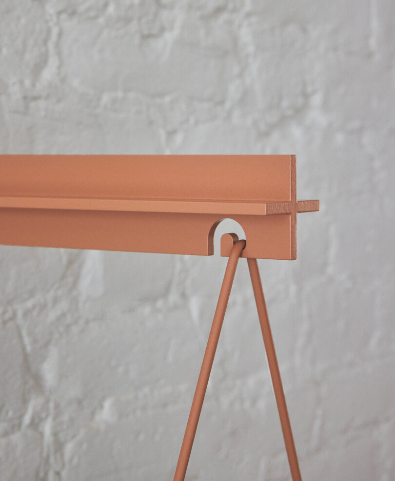 Close-up of a modern, minimalist metal structure painted in a reddish-orange color against a textured white brick wall. The structure has a triangular base and a horizontal bar featuring a small cutout.