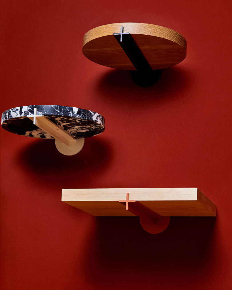 Three wall-mounted shelves against a red background, featuring distinct wood, marble, and metal finishings, each supported by unique, contrasting brackets.