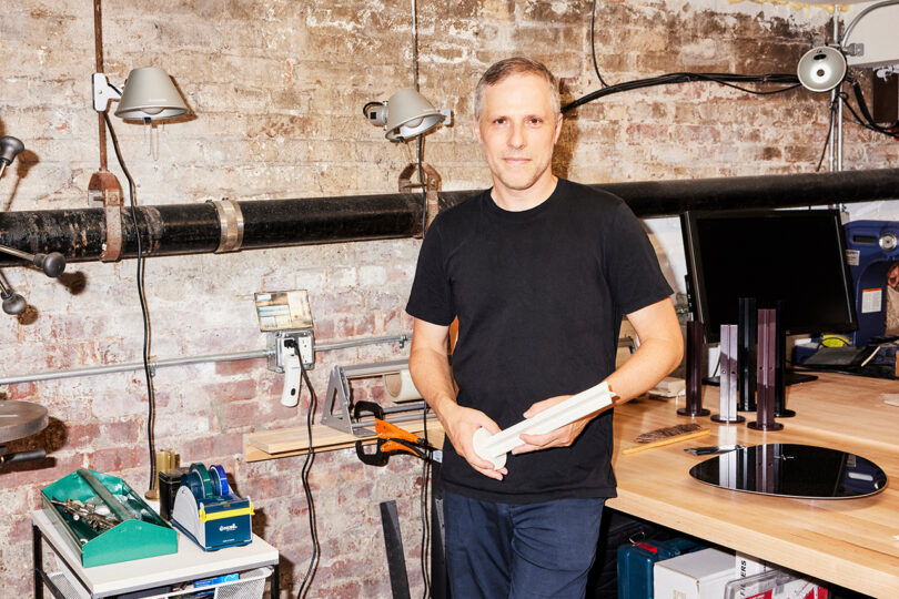 A man in a black T-shirt stands in a workshop holding a white object, with tools and machinery visible around him.