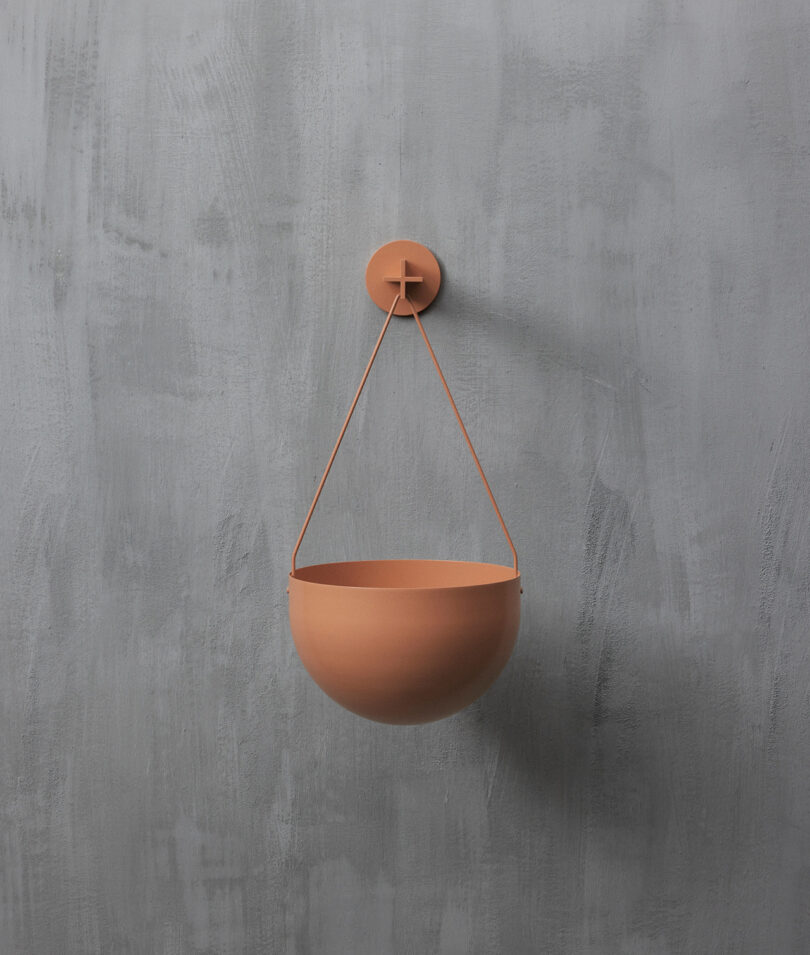 A round, terracotta-colored hanging planter is suspended on a grey textured wall by two thin straps attached to a circular wall hook.