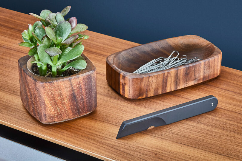 A small potted plant, a wooden tray with paperclips, and a black Task Knife by Grovemade are placed on a wooden surface.