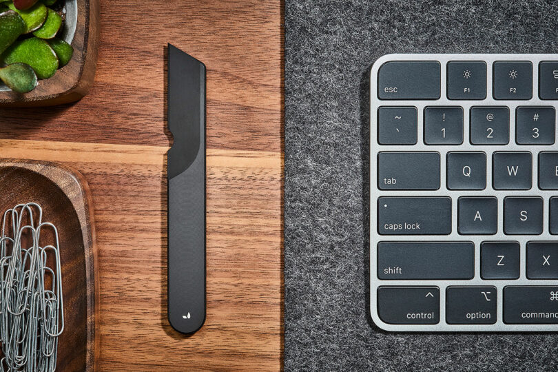 A desk with a close-up view of a keyboard, a black Task Knife from Grovemade, paper clips in a wooden tray, and a corner of a plant.